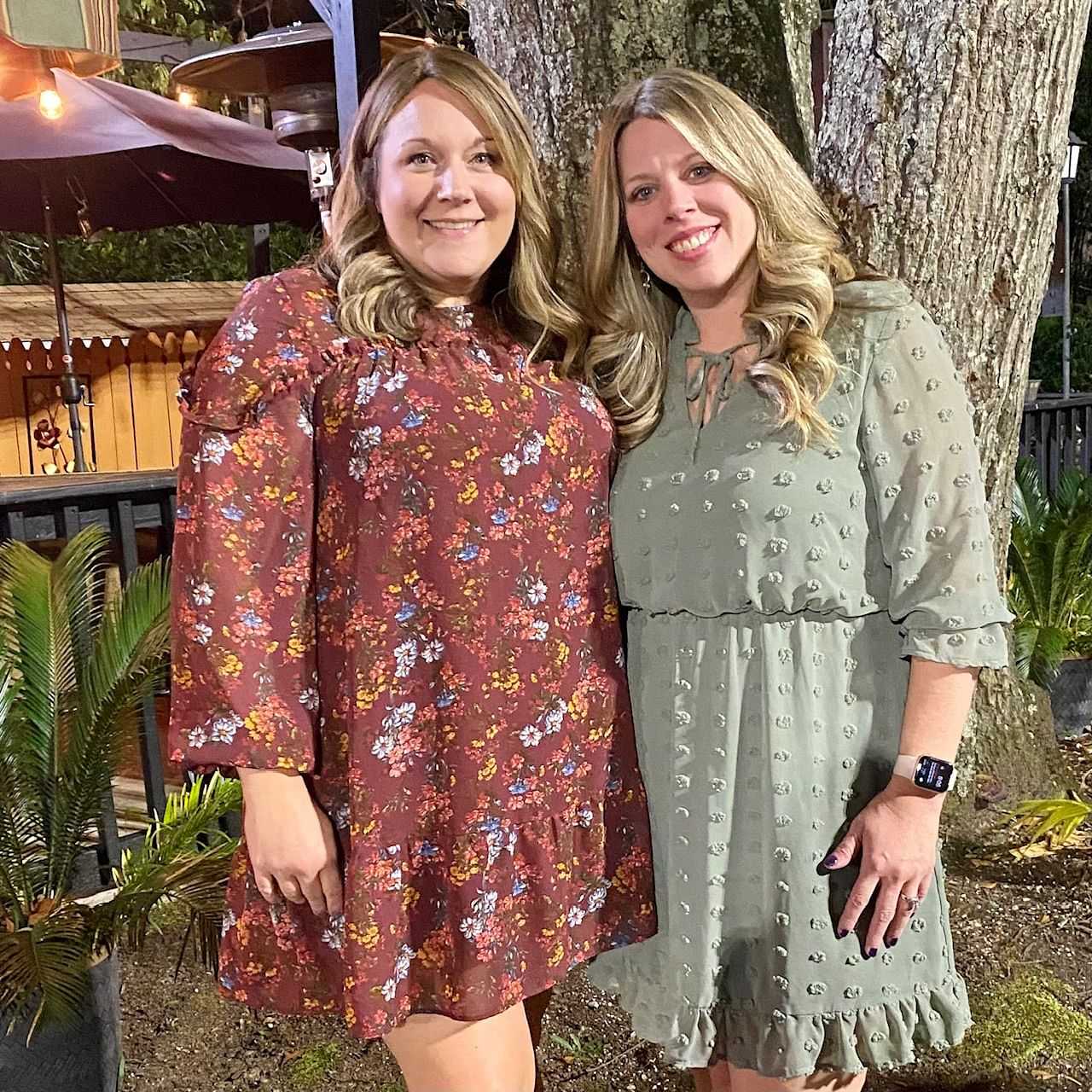 Two women smiling, standing outdoors at night, wearing floral and green dresses.